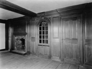 Black and white interior photo of a colonial-era study, showing a door, a glass-front hutch, and a fireplace
