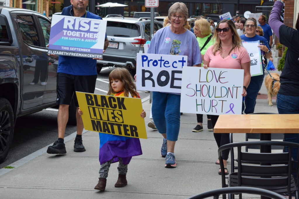 Young child wrapped in rainbow flag walks down Great Barrington sidewalk holding "Black Lives Matter" sign. Three adults follow holding signs saying "LOVE SHOULDN'T HURT", "Protect Roe", and "Rise Together for Safety & Justice"